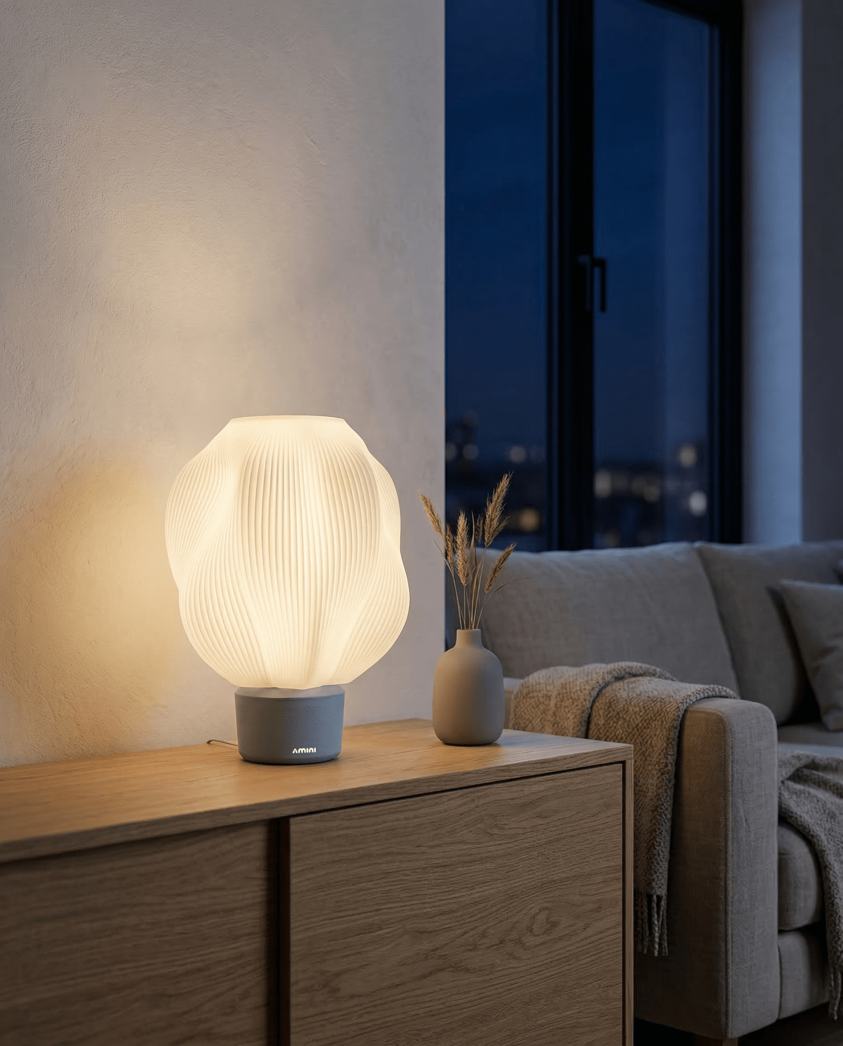 AMINI STRIA Lamp in Baby Blue — lit at night on a sideboard with dried grass vase, city lights visible outside